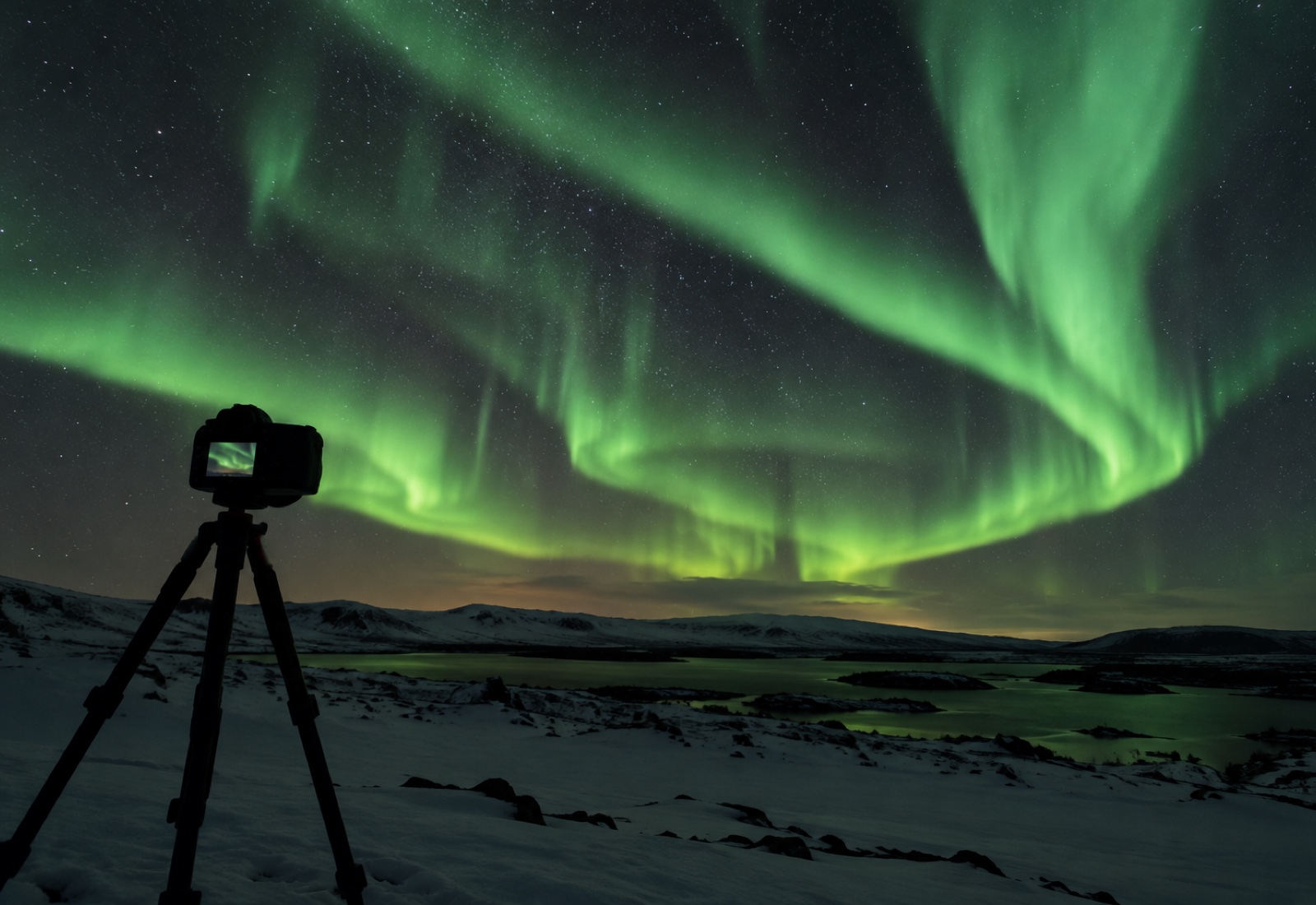 A camera on a tripod photographing an active green aurora display over a snowy landscape at night